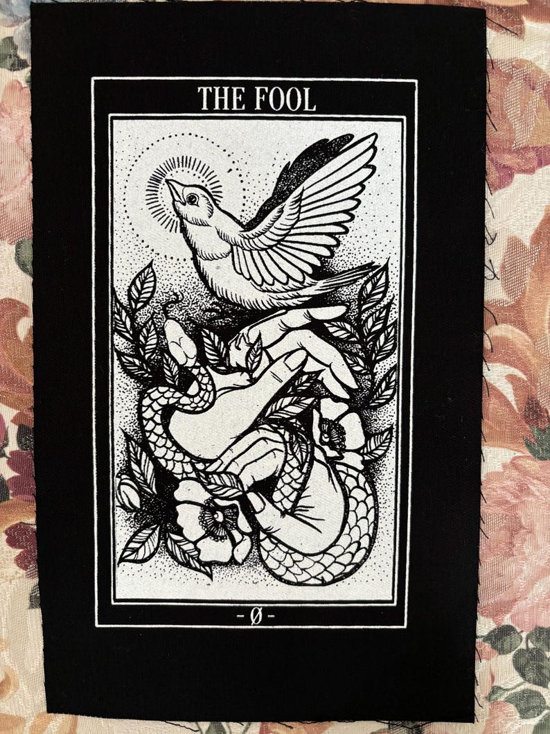 Op de afbeelding: Zwart-witte tarotkaart patch met de tekst "THE FOOL" bovenaan. De kaart toont een hand die een vogel en een slang vasthoudt met een bloem op de achtergrond.