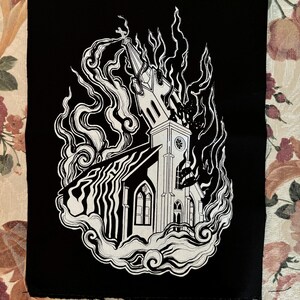 May include: Black and white embroidered patch of a church engulfed in flames. The church has a steeple and a clock tower. The flames are stylized and look like smoke.