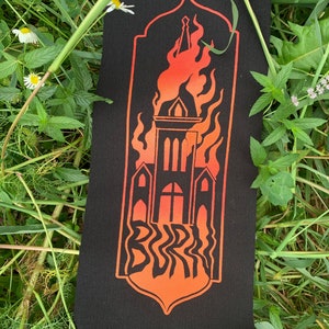 Pode incluir: Um remendo preto com um design degradê laranja e vermelho de um edifício de igreja envolto em chamas. A palavra "BURN" está escrita em uma fonte estilizada abaixo do edifício.