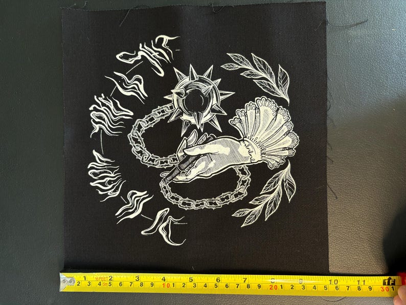 K&ouml;nnte beinhalten: Schwarzer quadratischer Aufn&auml;her mit wei&szlig;er Illustration. Das Design zeigt eine Hand, die eine Kette und eine Streitaxt h&auml;lt, umgeben von stilisiertem Text und Bl&auml;ttern. Der Aufn&auml;her misst ca. 10 cm x 10 cm.