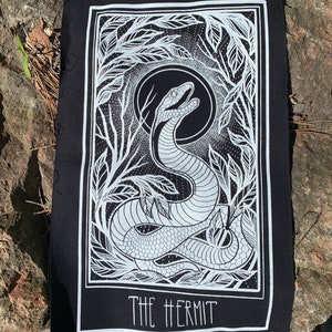 Op de afbeelding: Zwart-witte patch met een wit slangenontwerp en de tekst "THE HERMIT" in een wit rechthoek.