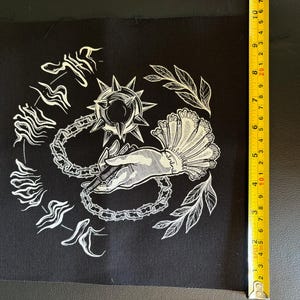 K&ouml;nnte beinhalten: Schwarzer quadratischer Aufn&auml;her mit wei&szlig;er Illustration. Das Design zeigt eine Hand, die eine Flegel h&auml;lt, umgeben von Kettengliedern, Bl&auml;ttern und stilisiertem Text. Der Aufn&auml;her ist etwa 18 cm hoch.