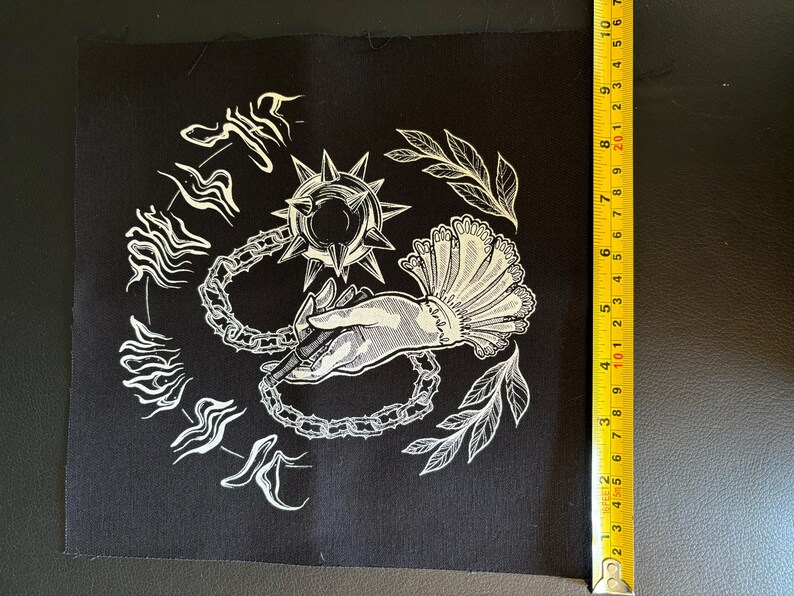 K&ouml;nnte beinhalten: Schwarzer quadratischer Aufn&auml;her mit wei&szlig;er Illustration. Das Design zeigt eine Hand, die eine Flegel h&auml;lt, umgeben von Kettengliedern, Bl&auml;ttern und stilisiertem Text. Der Aufn&auml;her ist etwa 18 cm hoch.