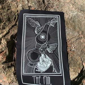 Op de afbeelding: Zwart-witte tarotkaart patch met het ontwerp van de kaart De Dwaas. De kaart toont een hand die een lantaarn vasthoudt met een mot die eromheen vliegt. De tekst "THE FOOL" is onderaan de kaart gedrukt.