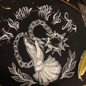 Op de afbeelding: Zwart vierkantige patch met een witte illustratie van een hand die een spijkerknuppel vasthoudt die met een ketting is verbonden. De hand draagt een gefranjerde manchet. Witte tekst in een cirkelvormig patroon omringt de centrale afbeelding. Witte blad- en abstracte ontwerpen zijn ook aanwezig.