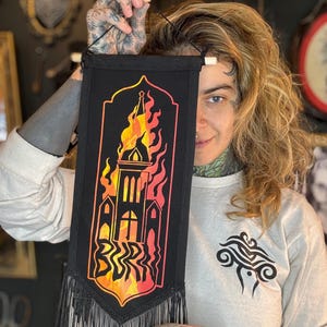 Può includere: Un arazzo da parete in tessuto nero con un'illustrazione in nero e arancione di un edificio avvolto dalle fiamme. L'edificio ha un tetto a punta e finestre. Le fiamme sono stilizzate e hanno un gradiente di arancione e giallo. Il tessuto ha un bordo a frangia.