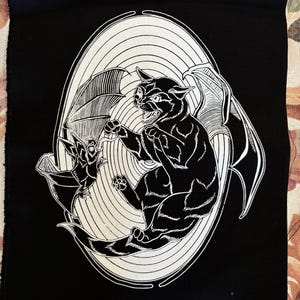 May include: Black and white illustration of a cat with bat wings attacking a bat. The image is on a black fabric background.