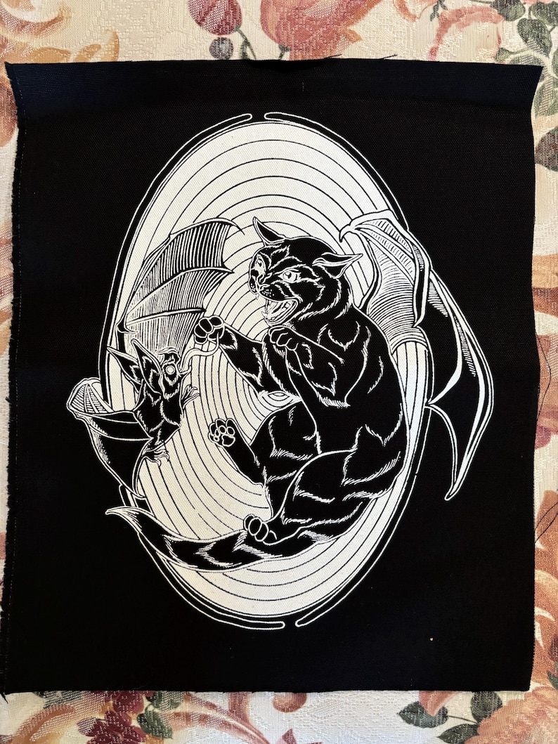 May include: Black and white illustration of a cat with bat wings attacking a bat. The image is on a black fabric background.