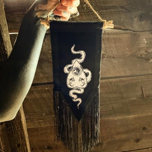 Puede incluir: Tapiz de pared de tela negra con un diseño blanco de una serpiente envuelta alrededor de un colgante triangular con un ojo en su interior. El tapiz tiene flecos negros alrededor de la parte inferior.