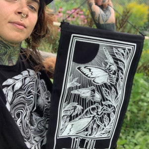 Op de afbeelding: Zwart-witte tarotkaart wandtapijt met de kaart van de Dwaas met een nachtvlinder en andere insecten. De kaart hangt aan een touwtje en heeft een zwarte achtergrond met witte tekst die "The Fool" luidt.