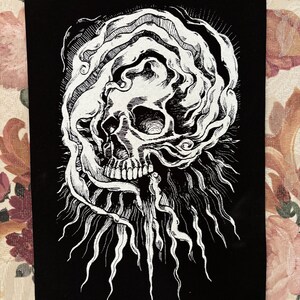 Op de afbeelding: Een geborduurde patch in zwart en wit met een schedel met een draaiende vorm eromheen. De schedel is gedetailleerd met tanden en een oogkas. Het ontwerp is omgeven door een stralend patroon van lijnen.