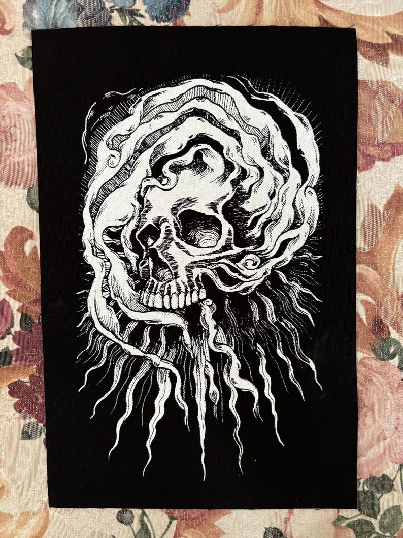 Op de afbeelding: Een geborduurde patch in zwart en wit met een schedel met een draaiende vorm eromheen. De schedel is gedetailleerd met tanden en een oogkas. Het ontwerp is omgeven door een stralend patroon van lijnen.