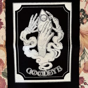 May include: Black and white embroidered patch with a skeletal hand holding a Ouija board planchette. The patch has a white border and the word "GOODBYE" is written below the image.