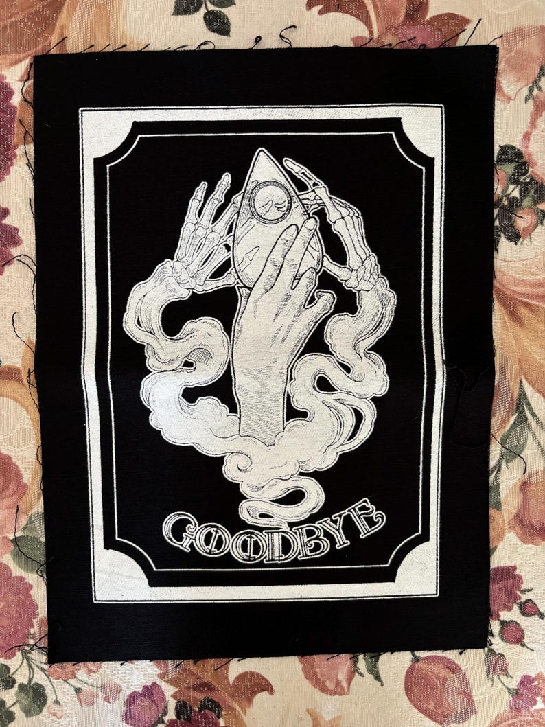 May include: Black and white embroidered patch with a skeletal hand holding a Ouija board planchette. The patch has a white border and the word "GOODBYE" is written below the image.