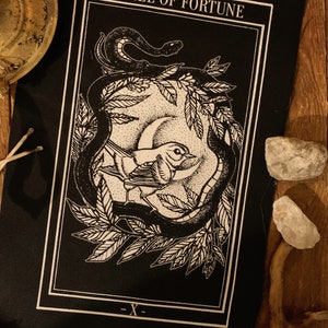 Op de afbeelding: Zwart-witte tarotkaart patch met het ontwerp van het Rad van Fortuin. De kaart toont een slang die om een ​​vogel is gewikkeld, met bladeren en takken eromheen. Het kaartnummer, X, is gedrukt in het midden onderaan.