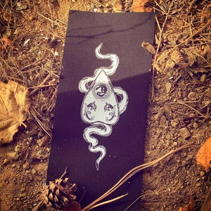 Puede incluir: Un parche de tela negro con un contorno blanco de una tabla Ouija con una serpiente envuelta alrededor. La serpiente tiene un solo ojo en la cabeza.