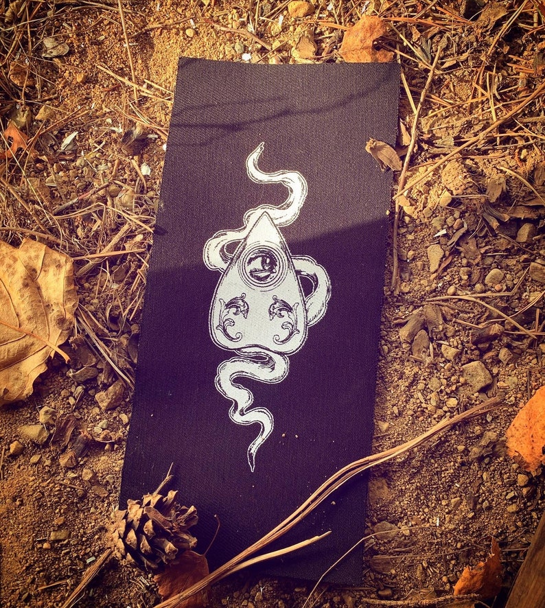 Puede incluir: Un parche de tela negro con un contorno blanco de una tabla Ouija con una serpiente envuelta alrededor. La serpiente tiene un solo ojo en la cabeza.