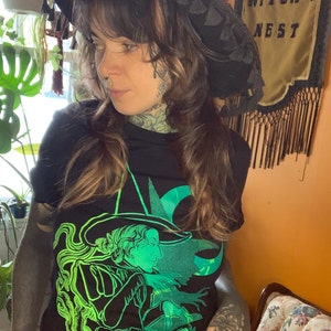Peut inclure: Un t-shirt noir avec un imprimé graphique vert d'une sorcière portant un chapeau et tenant un livre. La sorcière est entourée de nuages et d'étoiles. Le texte "The Witch's Nest" est visible en arrière-plan.