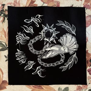 K&ouml;nnte beinhalten: Schwarzer quadratischer Aufn&auml;her mit einer wei&szlig;en Illustration. Das Design zeigt eine Stachelkugel, die durch eine Kette mit einer Hand verbunden ist, die einen Stift h&auml;lt, umgeben von stilisiertem Text und Bl&auml;ttern. Der Aufn&auml;her befindet sich auf einer Oberfl&auml;che mit floralem Muster.