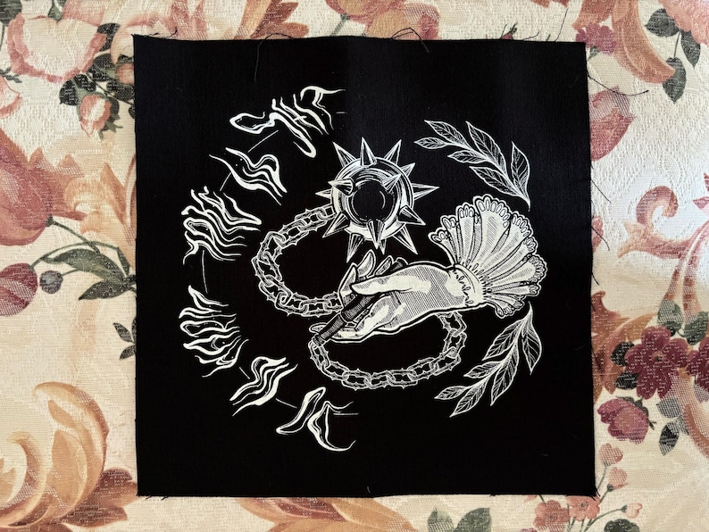 K&ouml;nnte beinhalten: Schwarzer quadratischer Aufn&auml;her mit einer wei&szlig;en Illustration. Das Design zeigt eine Stachelkugel, die durch eine Kette mit einer Hand verbunden ist, die einen Stift h&auml;lt, umgeben von stilisiertem Text und Bl&auml;ttern. Der Aufn&auml;her befindet sich auf einer Oberfl&auml;che mit floralem Muster.