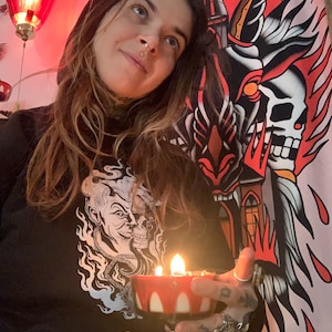 Pode incluir: Uma pessoa vestindo um moletom com capuz preto com um gráfico branco de um crânio e chamas. A pessoa está segurando uma vela vermelha e branca em um porta-velas de vidro.