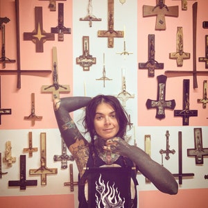 Peut inclure: Une personne avec des tatouages d'encre noire et un t-shirt noir avec une croix renversée blanche et des flammes dessus se tient devant un mur recouvert de croix renversées.