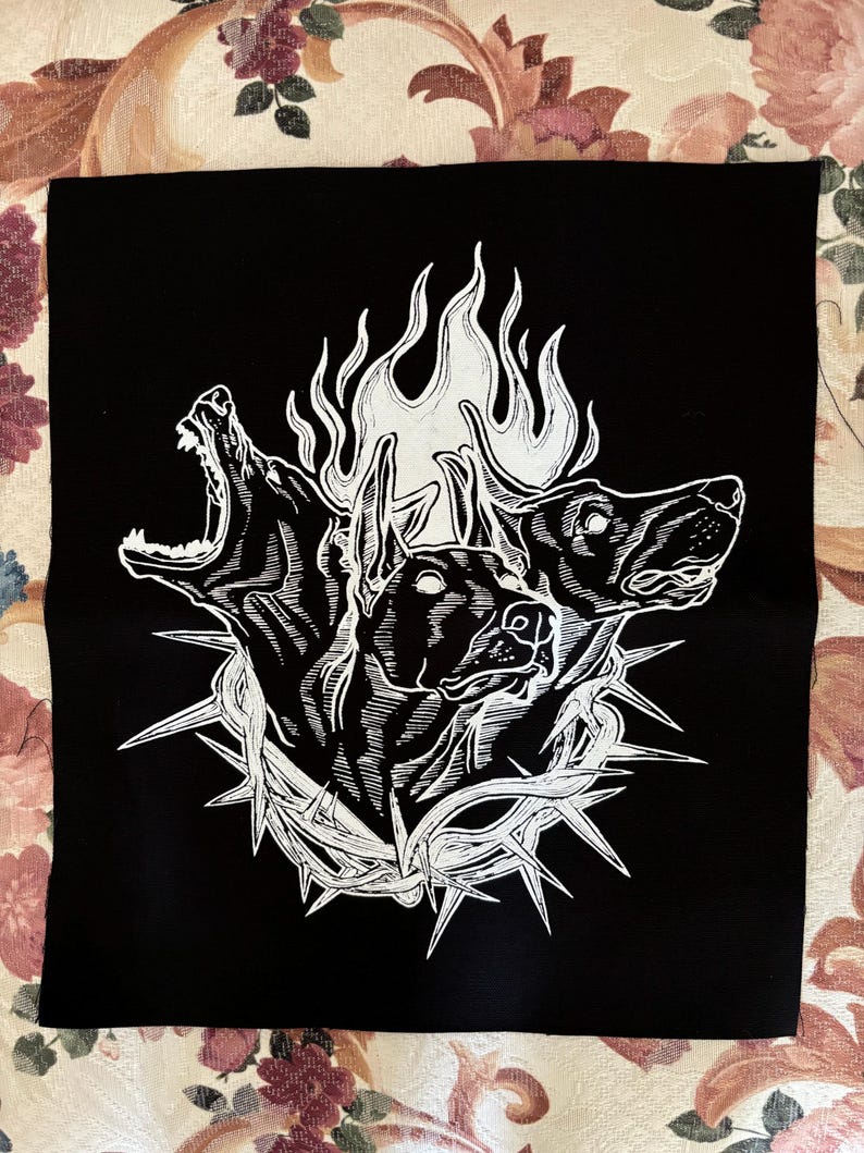 May include: Black and white patch with a graphic of two dog heads engulfed in flames and a crown of thorns.
