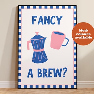 May include: A framed art print with a white background, featuring a blue coffee maker and a pink mug. The text "FANCY A BREW?" is in blue. A circular orange graphic states "More colours available". The frame has a blue and white checkered border.