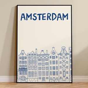 Puede incluir: Una impresi&oacute;n art&iacute;stica enmarcada con la palabra "AMSTERDAM" en azul sobre un dibujo lineal de casas de canal de &Aacute;msterdam. La obra de arte est&aacute; en un marco negro y tiene un fondo color crema.