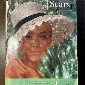 May include: Vintage Sears catalog cover featuring a woman wearing a white hat with a black band. The catalog is titled "Spring Through Summer 1964" and includes the Sears logo. The cover is aged with visible wear.