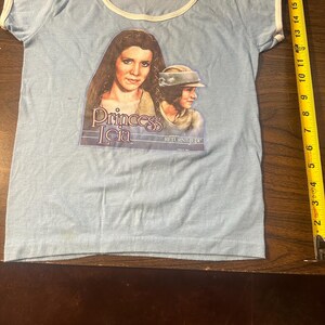 May include: Light blue t-shirt with white trim featuring a graphic of Princess Leia from Star Wars. The graphic includes the text "Princess Leia" and "RETURN JEDI". The shirt is a vintage item.