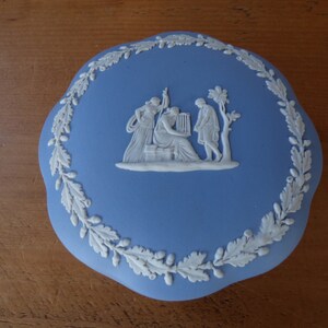 Op de afbeelding: Een blauwe en witte keramische sieradenbox met een reliëfdesign van drie figuren in een klassieke setting. De doos is versierd met een witte eikenbladerenrand.