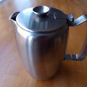 Puede incluir: Una cafetera de acero inoxidable con tapa y asa. La cafetera tiene un cuerpo redondeado y un pico para verter. La tapa tiene un pequeño pomo para facilitar la extracción. El asa tiene un diseño simple y curvado. La cafetera está sobre una superficie de madera.