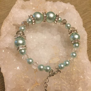 Puede incluir: Una pulsera de cuentas azul claro con detalles plateados. La pulsera presenta cuentas grandes y redondas y cuentas más pequeñas, con cuentas transparentes con forma de cristal. Los conectores y el cierre de metal plateado completan el diseño. La pulsera se muestra sobre una superficie blanca texturizada.