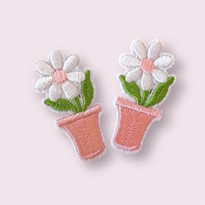May include: Two embroidered patches of a daisy in a pink flower pot. The daisies have white petals and a pink centre. Green leaves and stems are visible. The flower pots are a light pink colour.