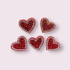 May include: Five red heart-shaped embellishments with a sparkling rhinestone border. The hearts are filled with small, red crystals. The embellishments are arranged on a light pink background.