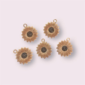 May include: Five gold-coloured sunflower charms with black centres. The charms are approximately 2.5 cm in diameter and have a loop at the top for attaching to jewellery or other crafts.