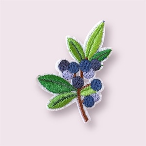May include: Embroidered patch depicting a blueberry sprig. The patch features green leaves with detailed stitching and a brown stem. The blueberries are a mix of blue and purple, with a white border. The patch is designed to be ironed on or sewn.