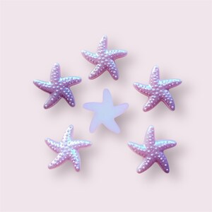 May include: Six iridescent starfish decorations in shades of pink and purple, arranged on a light pink background. Five of the starfish have a textured surface with small, raised dots. One starfish is a solid, lighter colour.