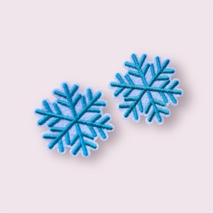 May include: Two embroidered snowflake patches. Each patch features a light blue centre with a darker blue snowflake design and a white border. The patches are set against a soft pink background.
