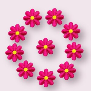 May include: Ten pink felt flower embellishments with yellow centers arranged in a circle on a white background.