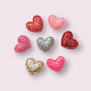 May include: A selection of glittery heart-shaped decorations in shades of red, pink, silver, and gold. The hearts are arranged on a pale pink surface. Each heart is covered in glitter, creating a sparkling effect.