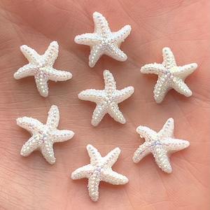 May include: Seven white, iridescent starfish decorations. Each starfish has five points and is covered in small, sparkling crystals. The decorations are arranged on a light-coloured surface, suggesting a beach or ocean theme.
