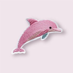 May include: Embroidered patch of a pink dolphin leaping. The dolphin has a white outline and a small black eye. The patch is made of pink and light pink thread with a small blue fin.