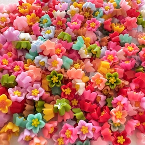 May include: A large assortment of colourful plastic flower-shaped embellishments. The flowers are in various shades of pink, blue, green, yellow, and red.