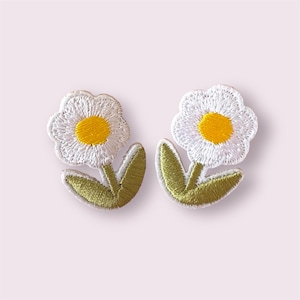 May include: Two embroidered daisy patches. Each patch features a white flower with a yellow centre, green stem, and green leaves. The patches are designed to be sewn onto fabric.