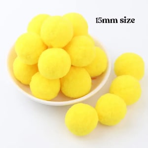 May include: A close-up of a pile of 15mm yellow pom poms. The pom poms are soft and fluffy, and they are perfect for crafting projects.