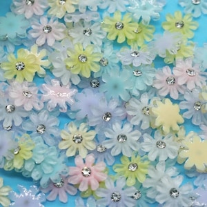 May include: Assortment of pastel coloured plastic flowers with clear rhinestone centres. The flowers are scattered on a blue background.