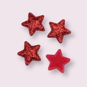 May include: Four red star-shaped embellishments. Three stars have a glittery texture, while the fourth has a solid, stitched appearance. The stars are arranged against a light pink background, creating a festive and decorative theme.