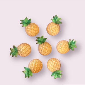 May include: Eight yellow pineapple-shaped buttons with green leaves. The buttons are arranged in a starburst pattern on a light purple background.
